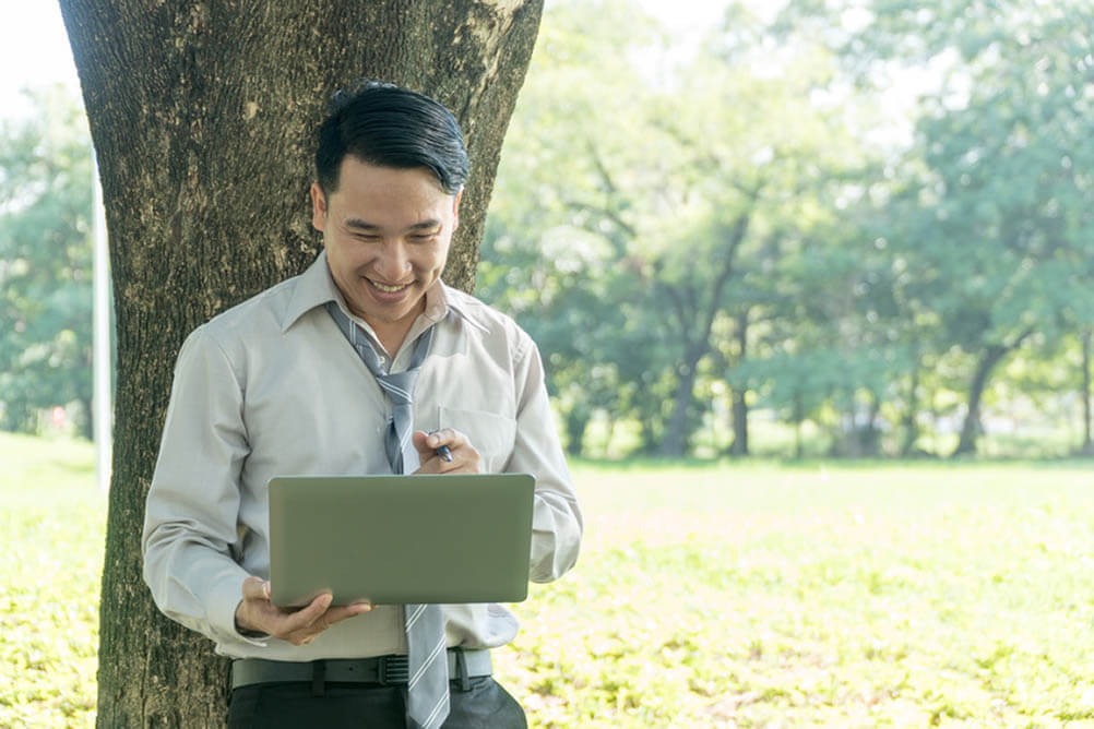 Man leans against tree holding laptop and smiling
